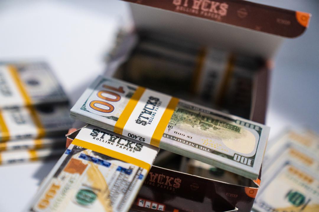 Close-up photo of stacks of $100 US dollar bills wrapped in yellow paper bands, sitting on what appears to be brown cardboard boxes. The image has a shallow depth of field with parts of the money in sharp focus while other areas are blurred